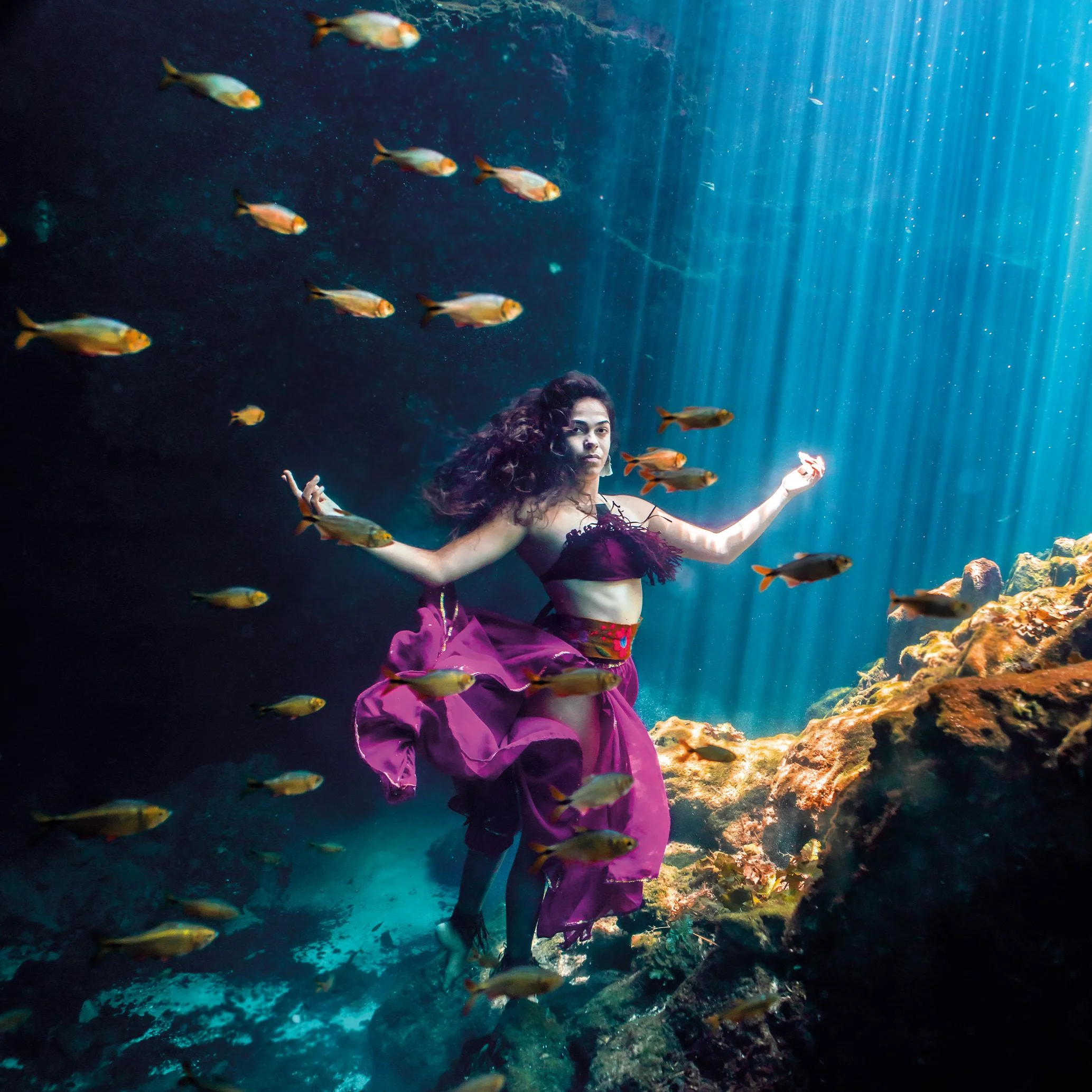 Figure in a magenta dress among fish in a shaft of cenote light
